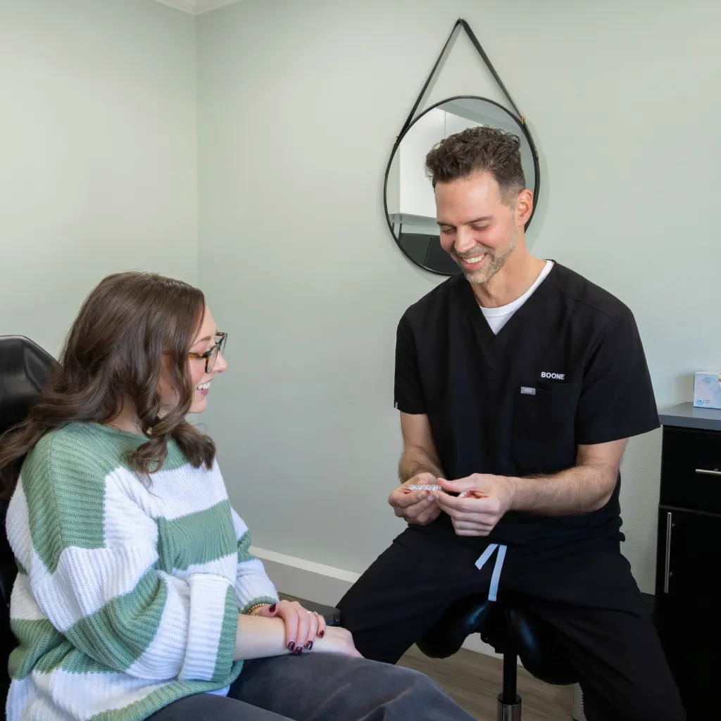 Happy woman with long hair holding a clear dental aligner, smiling and wearing a yellow shirt indoors.