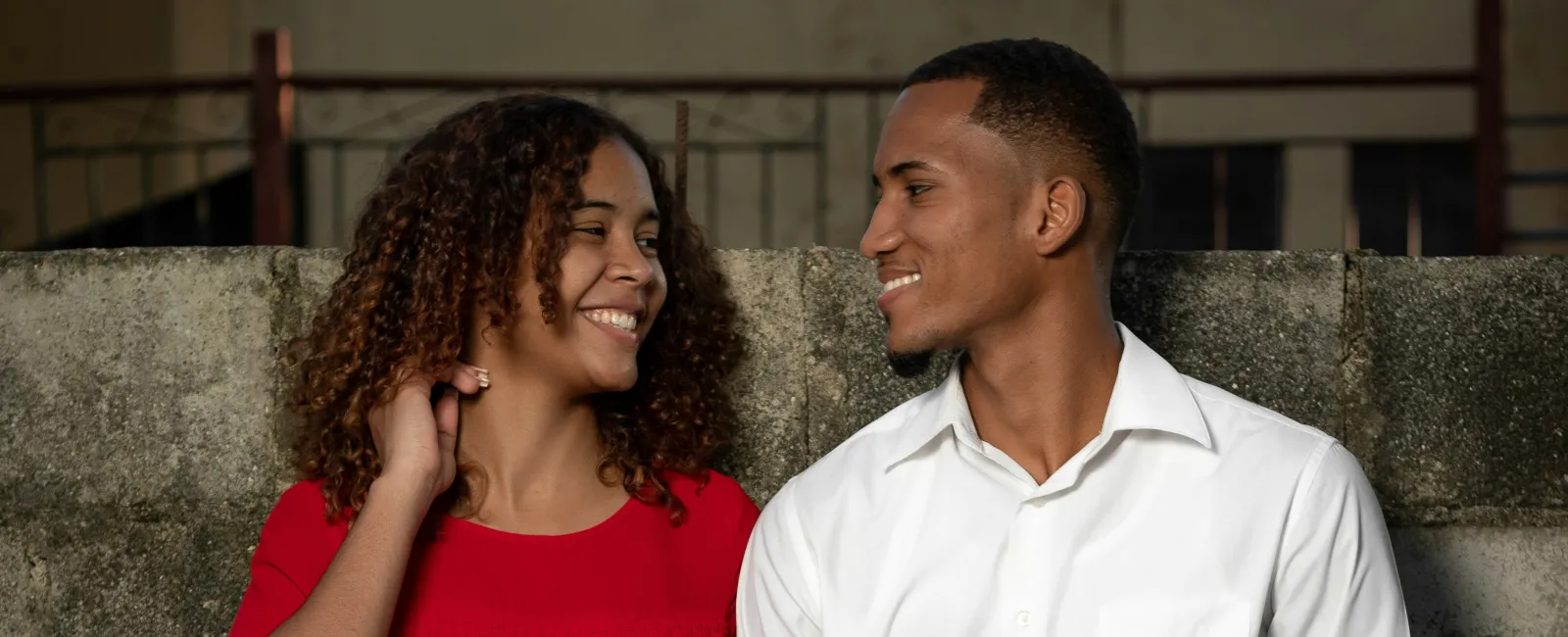 Smiling couple standing by a concrete wall holding hands, the woman in red blouse and the man in white shirt.