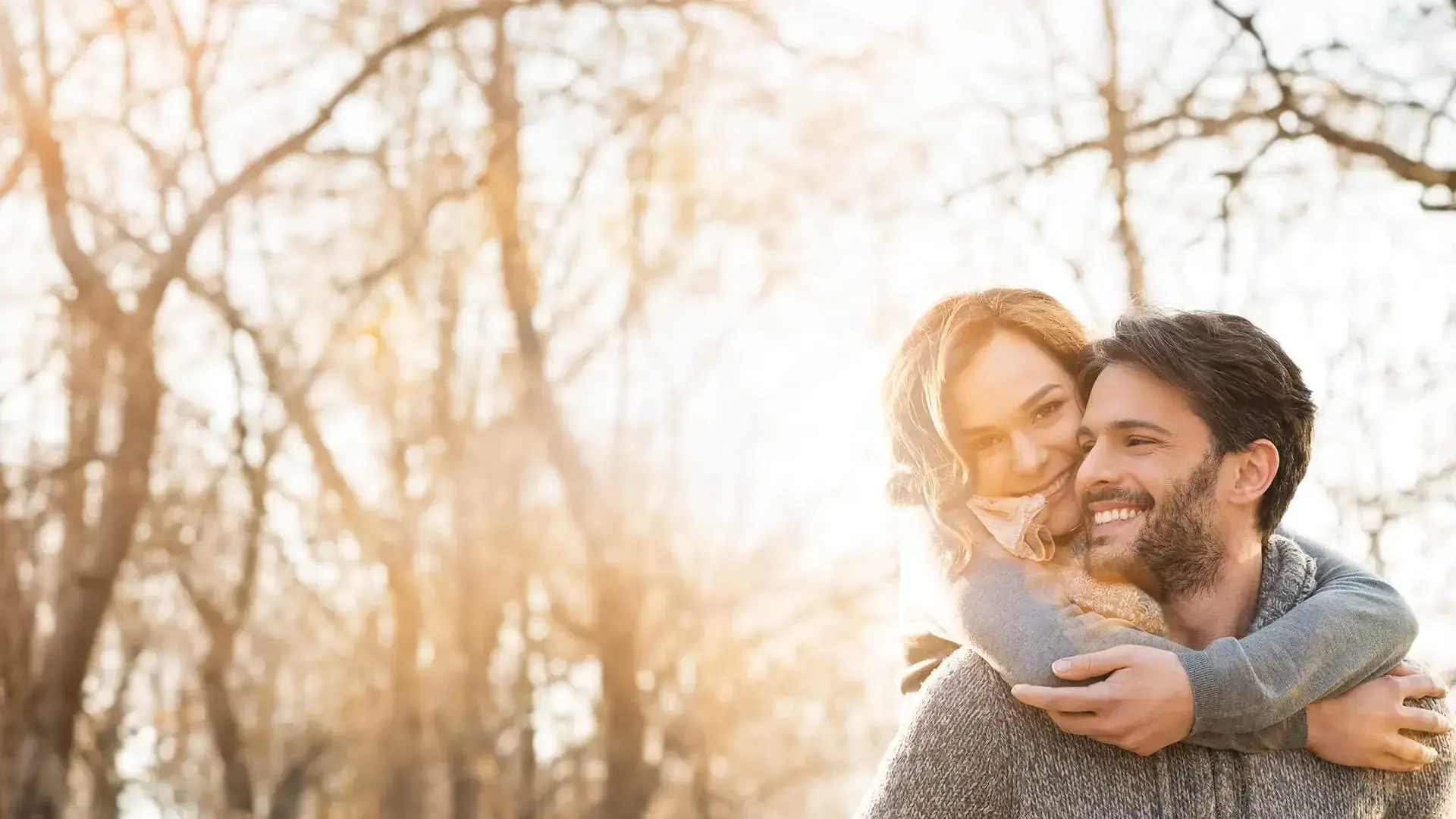 Happy couple outdoors in autumn with bare trees and warm sunlight, woman hugging man from behind smiling