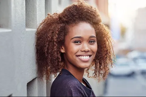 Smiling young woman with curly hair leaning against a wall in an outdoor urban setting with soft natural light.