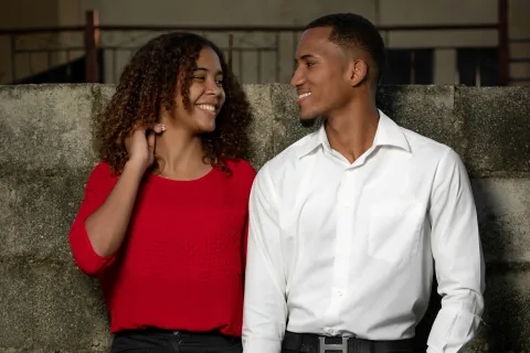 Smiling couple standing by a concrete wall holding hands, the woman in red blouse and the man in white shirt.