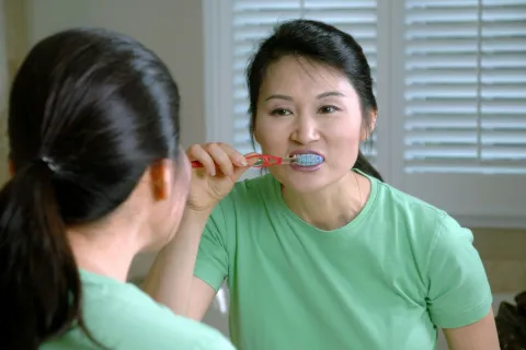 Woman wearing green shirt brushing teeth with a blue toothbrush in front of bathroom mirror.