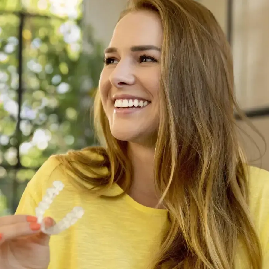 Happy woman with long hair holding a clear dental aligner, smiling and wearing a yellow shirt indoors.