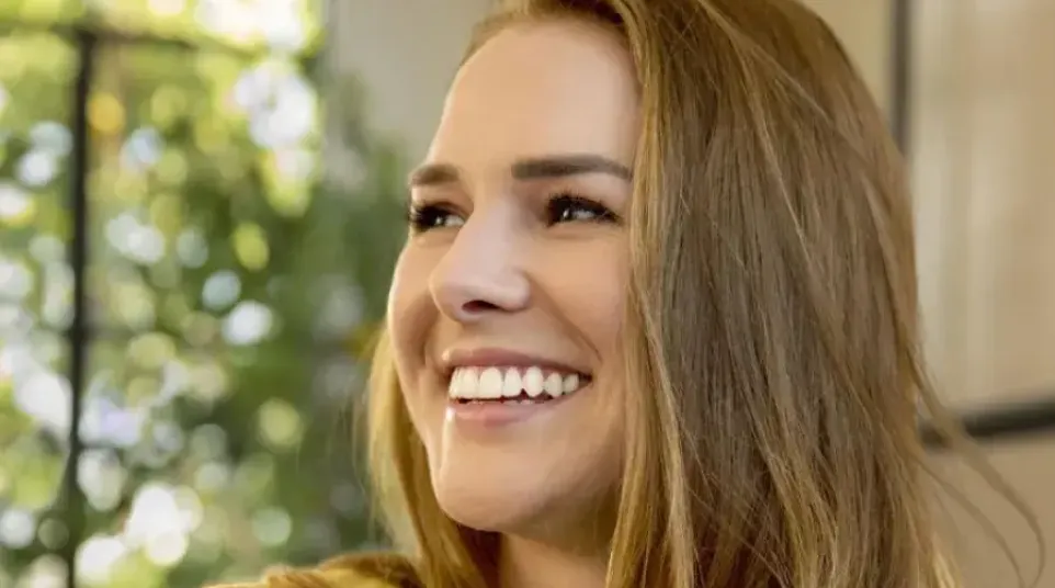 Happy woman with long hair holding a clear dental aligner, smiling and wearing a yellow shirt indoors.