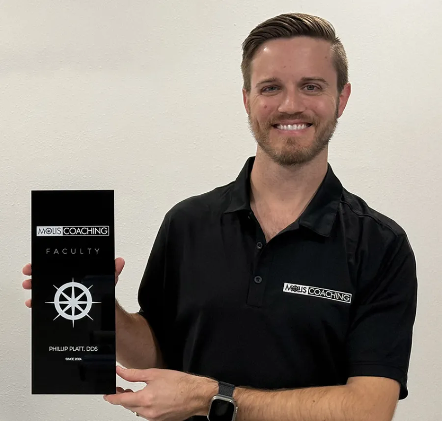 Smiling man in black Molis Coaching shirt holding a black faculty plaque with a compass logo.