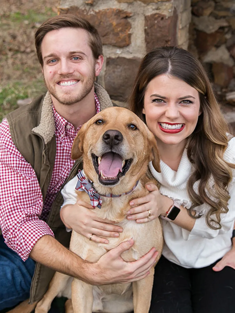 Happy couple sitting outdoors hugging a smiling golden retriever dog with a stone wall in the background