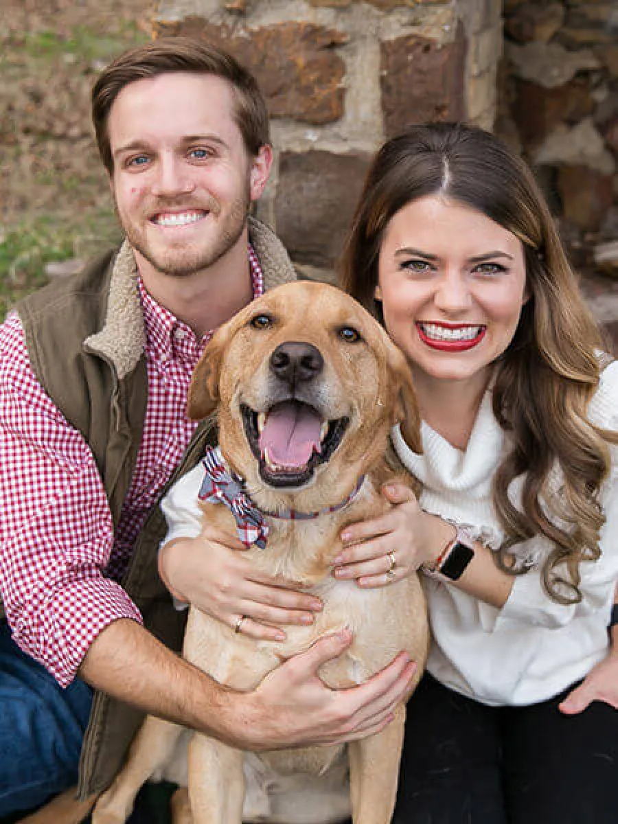 Smiling couple hugging a happy yellow Labrador dog outdoors in front of a stone wall