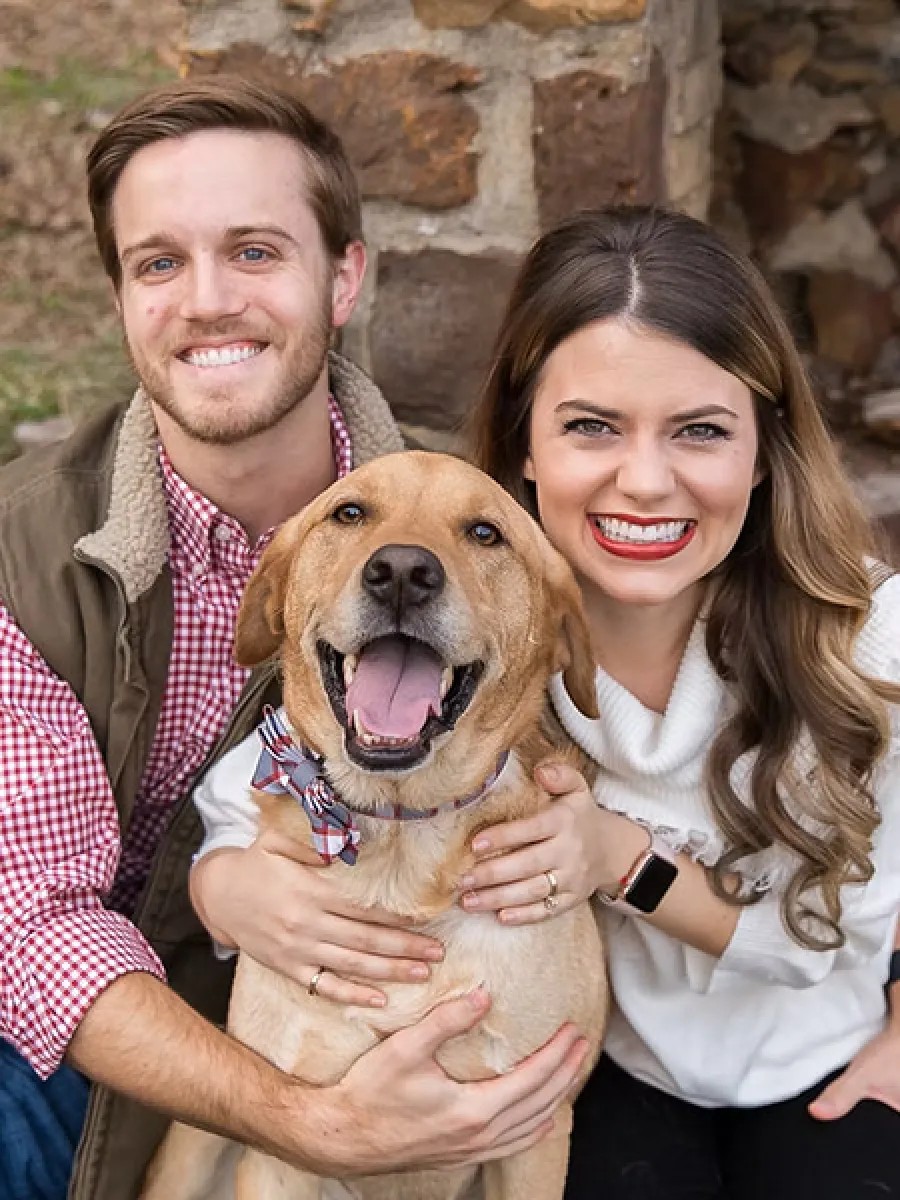Smiling couple embraces happy golden retriever wearing a bow tie outdoors by a stone wall